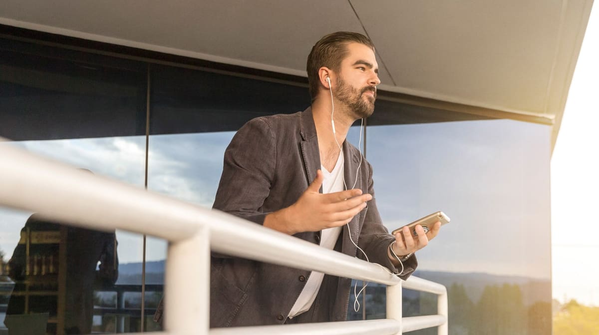 A man on a call through wired earbuds, phone in hand, leaning on a balcony rail outside an office