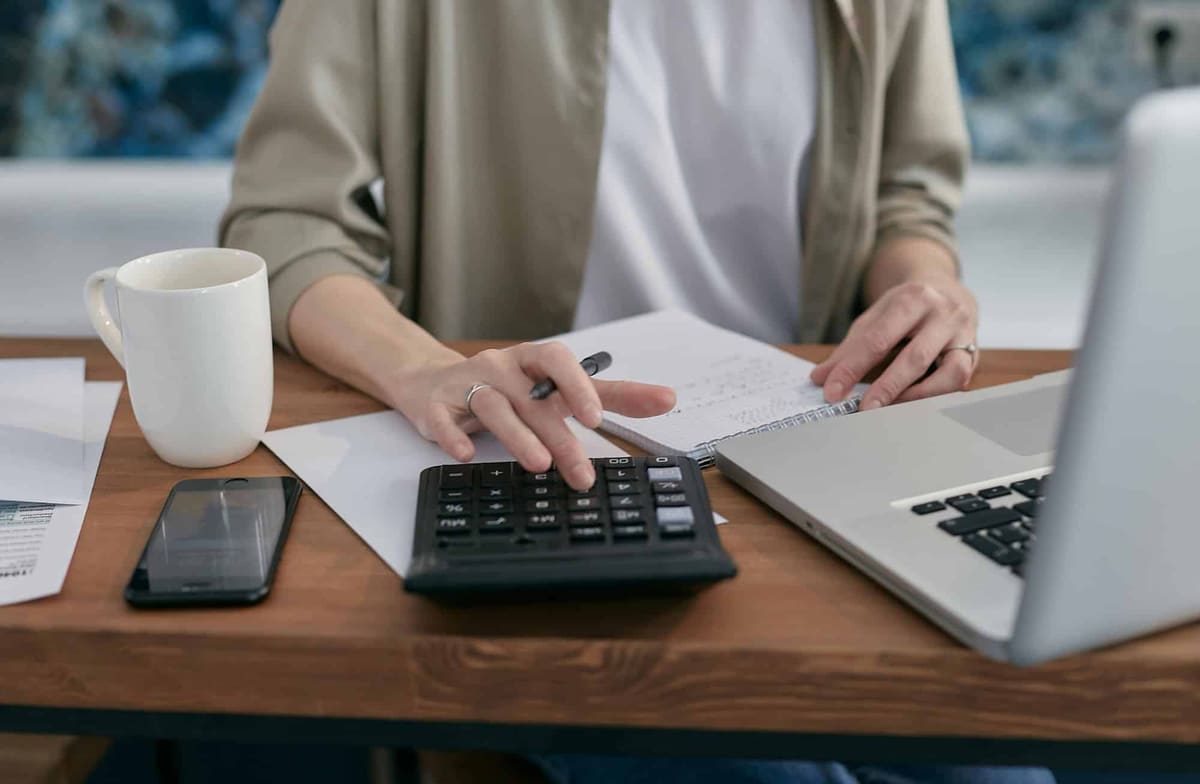 A person working a calculator at a wooden desk beside a notebook, a laptop, a phone and a coffee mug