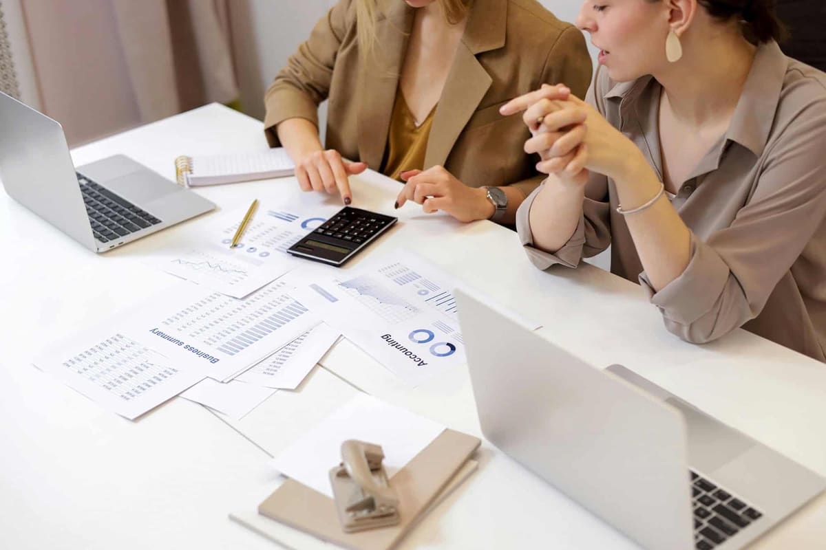 Two women going over printed reports and a calculator at a white desk with laptops