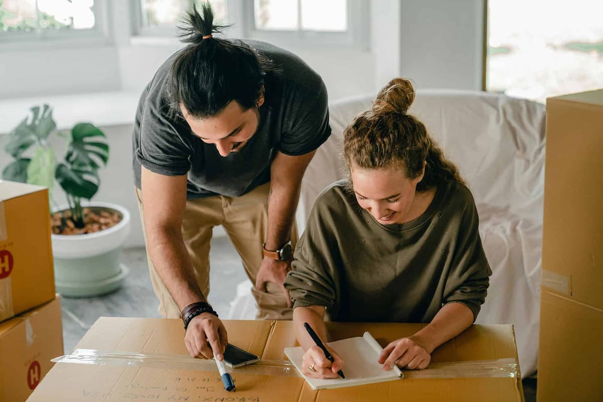 Two people at a taped moving box, one pointing with a marker while the other writes in a notebook