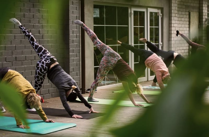 A yoga class holding a three-legged downward dog on mats on a wooden deck outside a brick studio