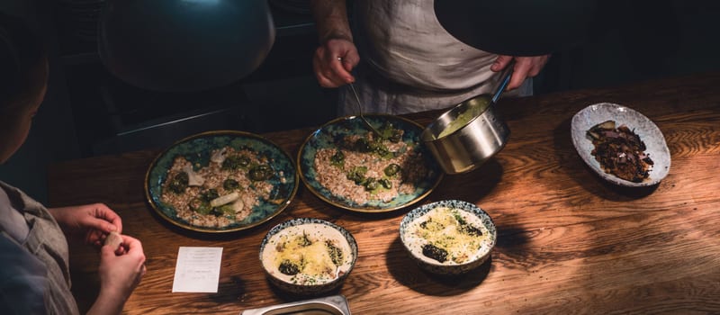 A chef plating four dishes on a wooden pass in an open restaurant kitchen