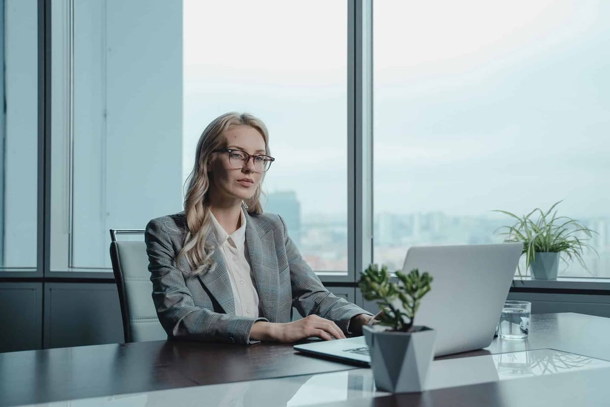 A woman in a gray blazer working at a laptop by a tall office window