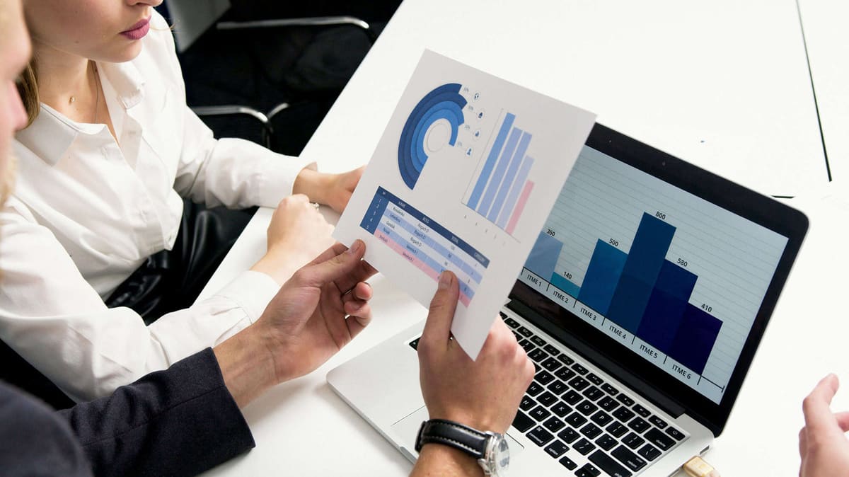 Two people comparing a printed chart with a bar chart on an open laptop