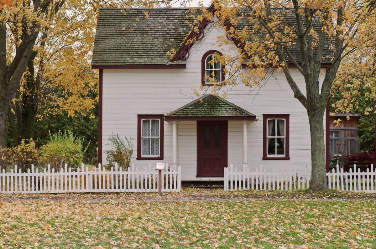 A white clapboard house with a red door, a shingle roof and a picket fence, autumn leaves on the lawn